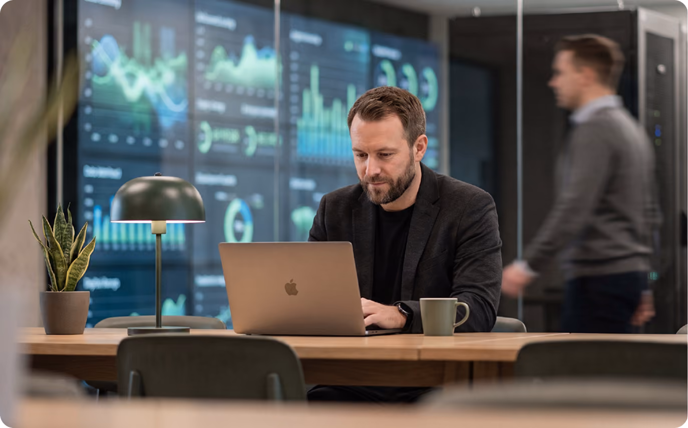 Man with beard working on a laptop at a wooden table with a cup and a plant, with large digital data charts displayed on glass wall behind him, and another man walking blurred in the background.