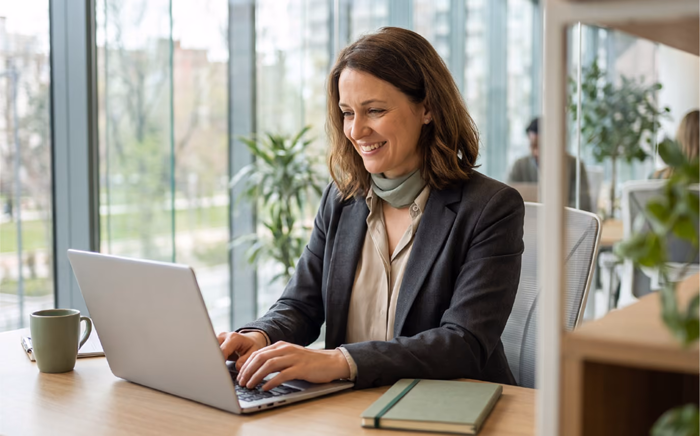 A smiling woman with brown hair wearing a dark blazer and light scarf types on a laptop at a wooden desk in a bright office with large windows and plants.