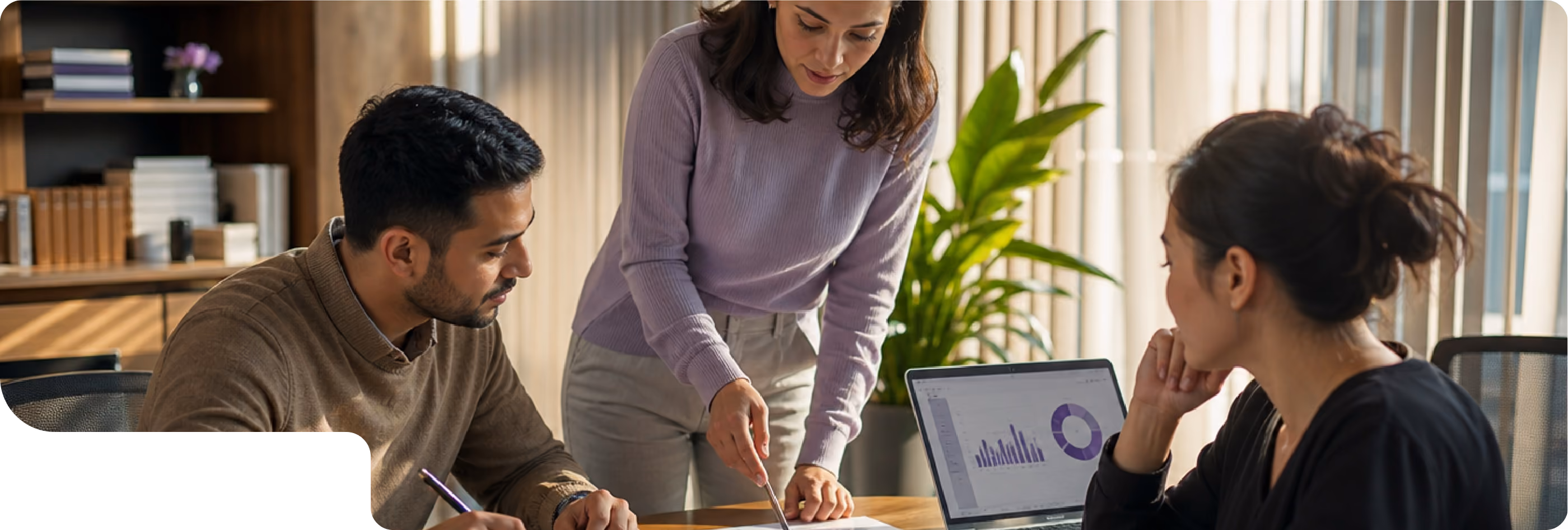 Three colleagues in an office discussing data shown on a laptop; a woman in a purple sweater points at a document while two others listen attentively.