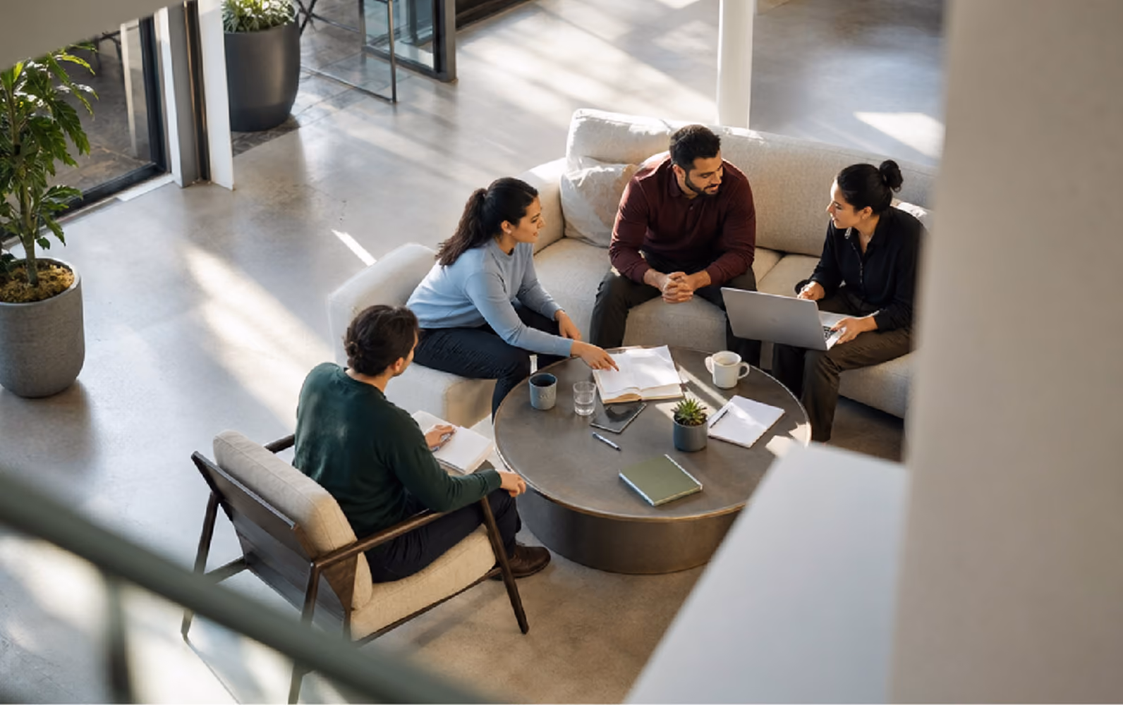 Four colleagues sitting around a round coffee table in a modern office lounge, engaged in a discussion with documents, notebooks, a laptop, coffee mugs, and a small plant on the table.