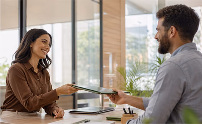 A woman and a man sitting at a desk in a modern office exchanging a folder while smiling.
