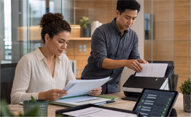 A woman sitting and reviewing documents at a desk while a man stands beside her scanning papers using a scanner in an office setting.