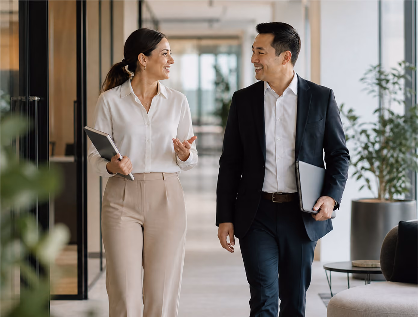 A man and a woman in professional attire smiling and walking together in a modern office hallway, each holding a laptop or folder.