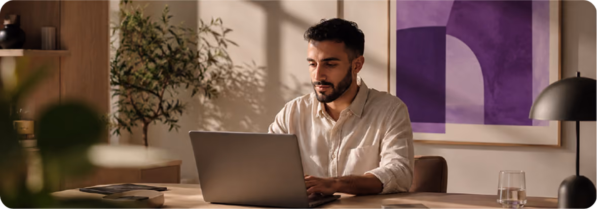 Bearded man in a beige shirt working on a laptop at a wooden desk in a warmly lit room with a plant, abstract purple wall art, and a glass of water.