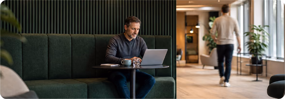 Man sitting on a green cushioned bench in a modern office lounge area, working on a laptop at a small table with a coffee cup and glass of water, while another person walks in the background near large windows.