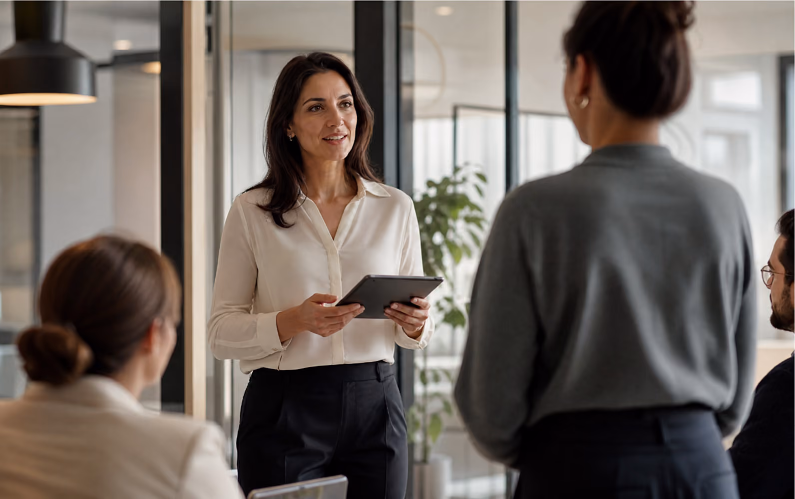 A professional woman wearing a white blouse holds a tablet and speaks to three colleagues in an office setting with glass walls and modern decor.