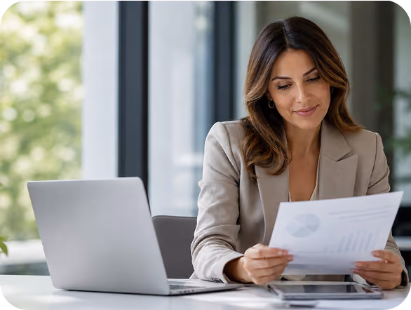 Businesswoman in a beige blazer sitting at a desk with an open laptop, reviewing a paper with charts in a modern office.