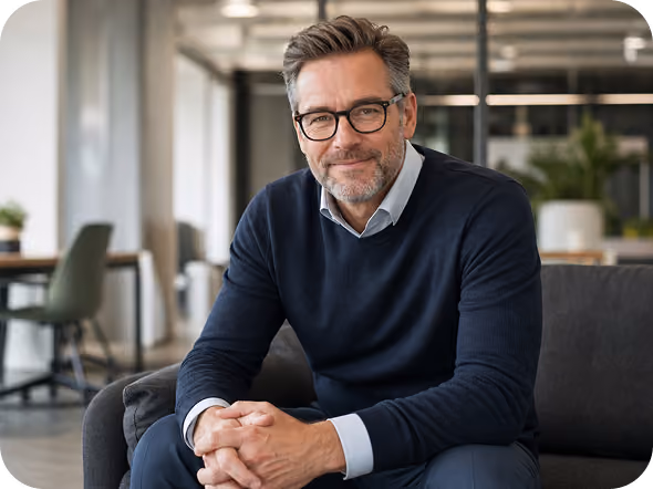 Middle-aged man with gray hair and glasses wearing a navy sweater and light blue shirt sitting on a dark sofa in a modern office setting, smiling and looking at the camera.
