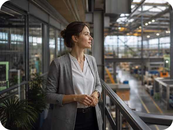 A professional woman with dark hair tied back, wearing a gray blazer and white blouse, standing indoors and looking thoughtfully out over a large industrial workspace with machinery and equipment.