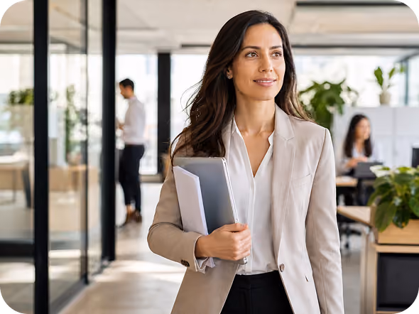Confident businesswoman in a beige blazer walks through a modern office holding a laptop and documents.