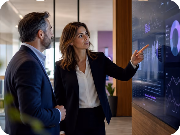 A woman in business attire points to data visualizations on a large screen while talking to a man in a suit in a modern office setting.
