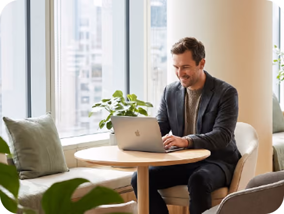 Man in a dark blazer sitting at a round wooden table near a window working on a laptop with a plant on the table and cushions on a bench nearby.