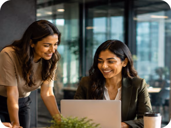 Two women smiling as they look at a laptop together in an office setting, with a coffee cup and plant nearby.