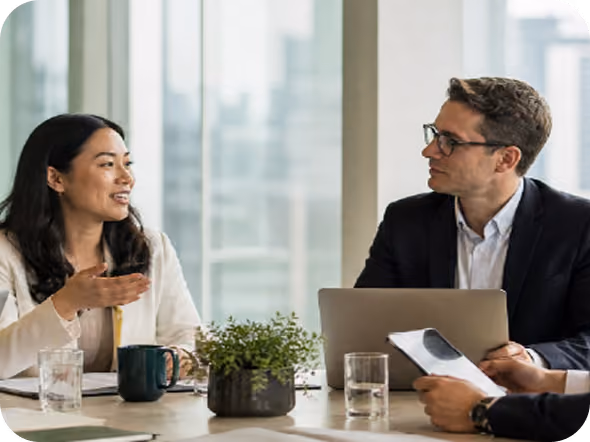 Two professionals, a woman and a man, engaged in a discussion at a table with a laptop, documents, glasses of water, a coffee mug, and a small potted plant in a modern office with large windows.