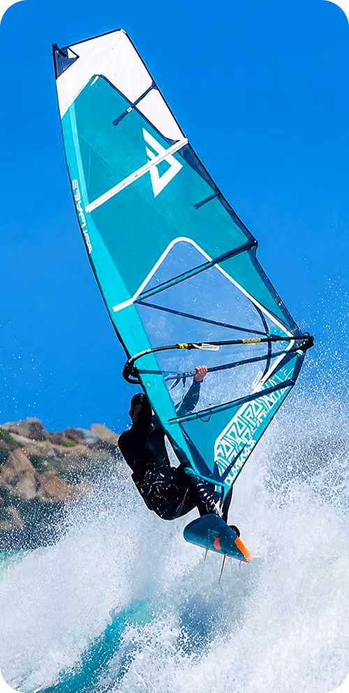 A windsurfer in a black wetsuit riding a turquoise and white sailboard over white ocean waves with a clear blue sky and rocky shoreline in the background.