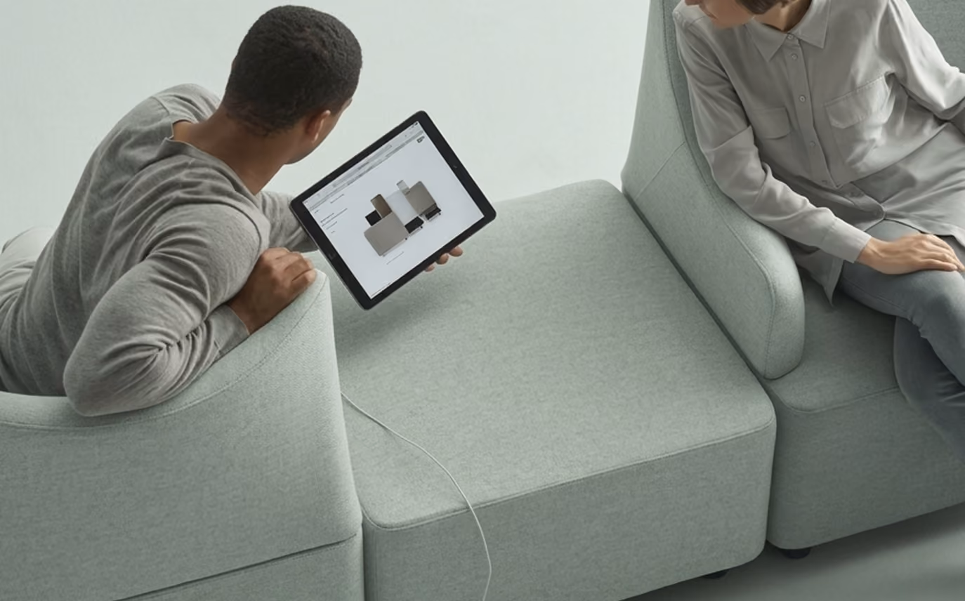 Two people sitting on a modular light gray sofa, one person holding a tablet displaying a 3D model or design, connected by a visible cable.