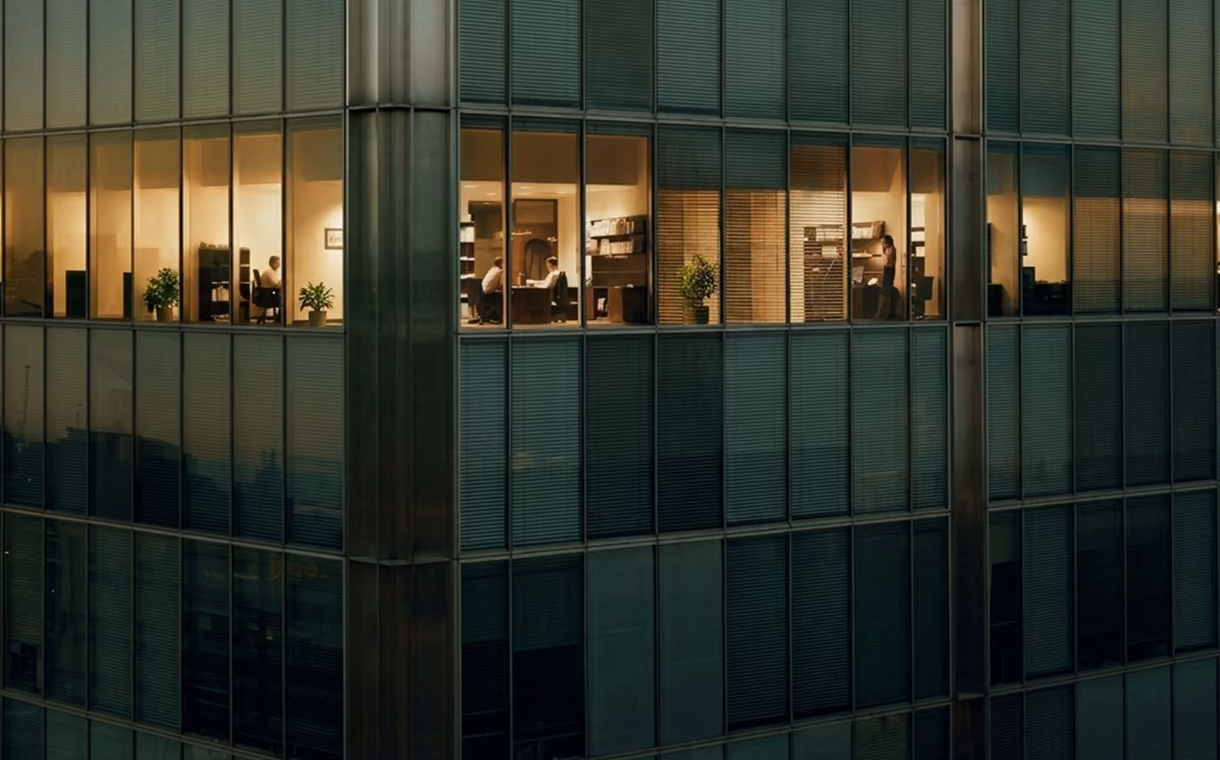 View of a modern office building facade at night showing three offices with illuminated windows where people are working and shelves with plants and books are visible.