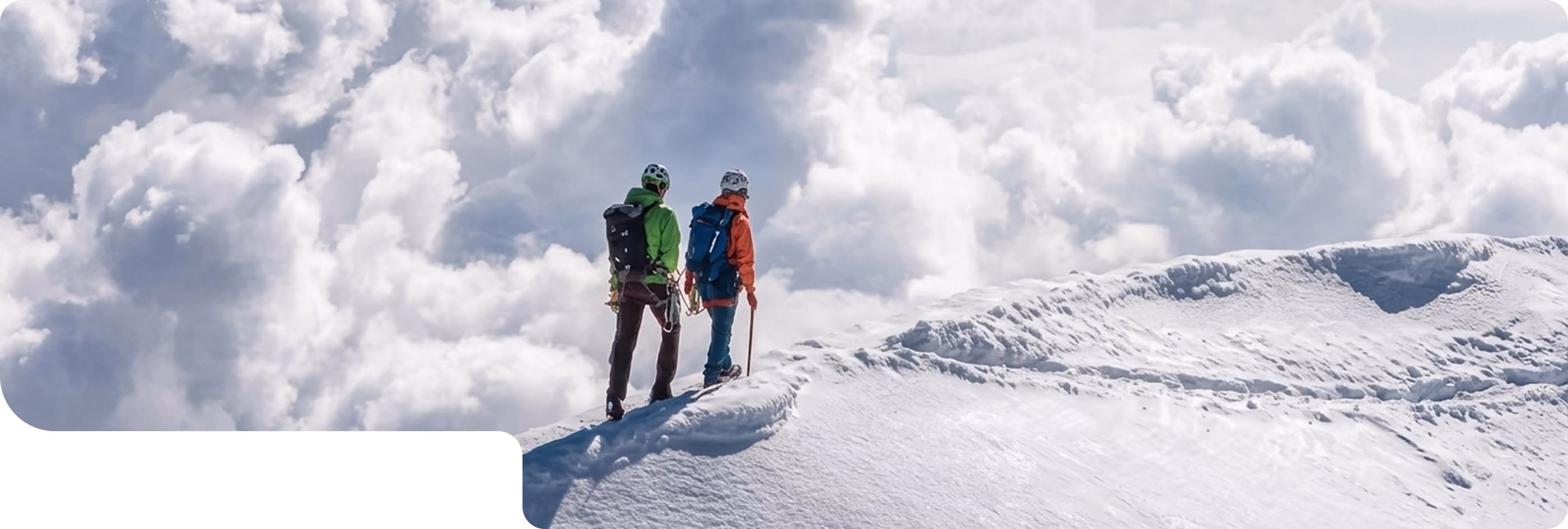 Two climbers wearing helmets and backpacks stand on a snowy mountain ridge above clouds.