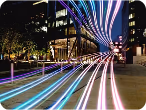 Nighttime urban scene with colorful light trails from moving vehicles along a city street flanked by modern buildings and trees.