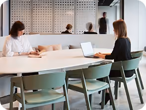 Two women seated at a modern white table in a minimalist office setting; one is reading a book and the other is typing on a laptop while glass walls reflect blurred figures walking by.