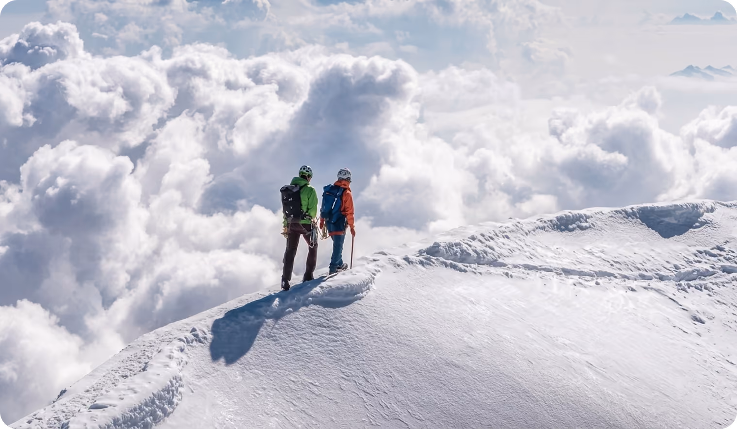 Two climbers standing on a snowy mountain ridge above thick clouds, wearing helmets and backpacks, one holding an ice axe.