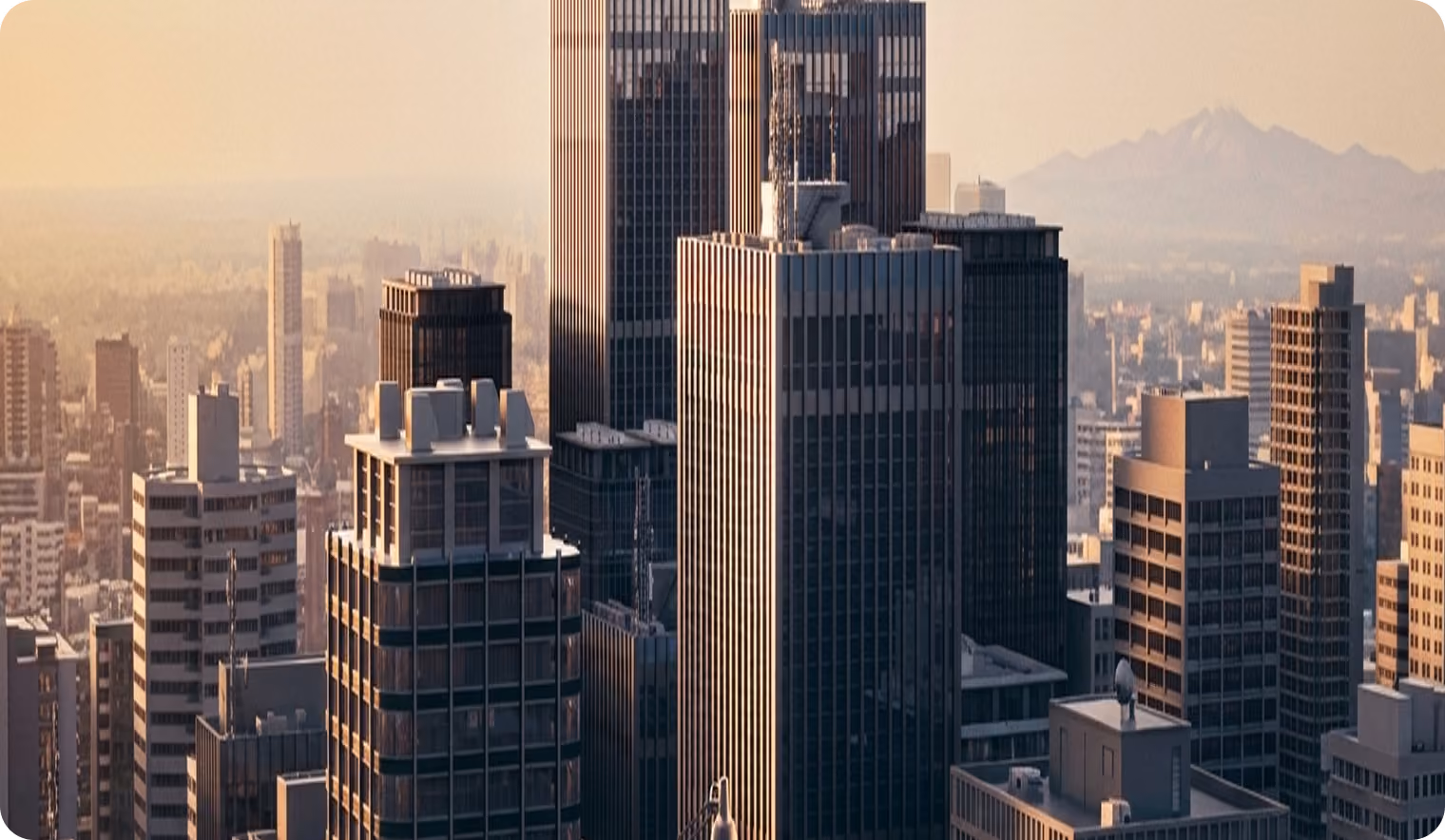 Cityscape at sunset featuring tall modern skyscrapers with soft golden light and a hazy distant mountain range in the background.