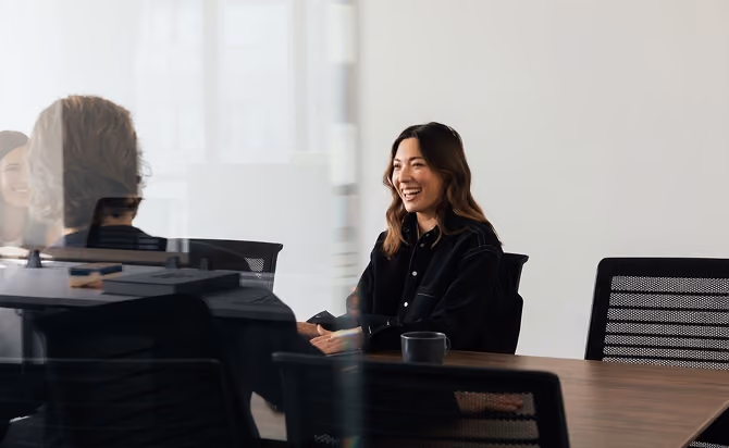 A woman with long brown hair, wearing a black shirt, smiling while seated at a conference table during a meeting, with office chairs and a coffee mug on the table.