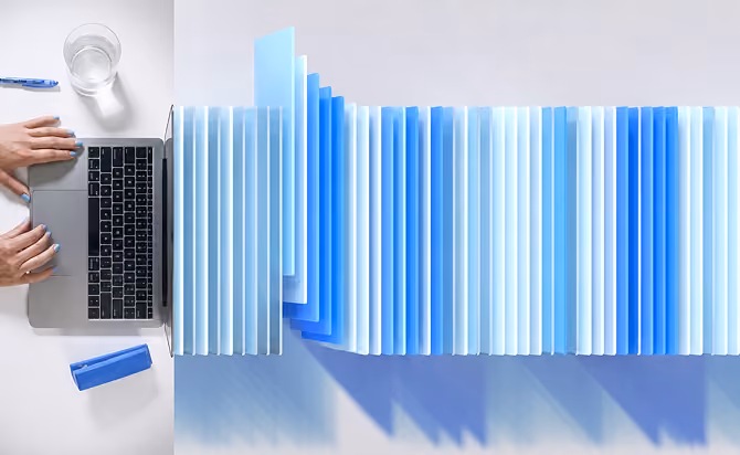 Overhead view of a laptop on a white desk with hands typing, next to a long row of blue file folders fanned out horizontally, a glass of water, and a blue pen.