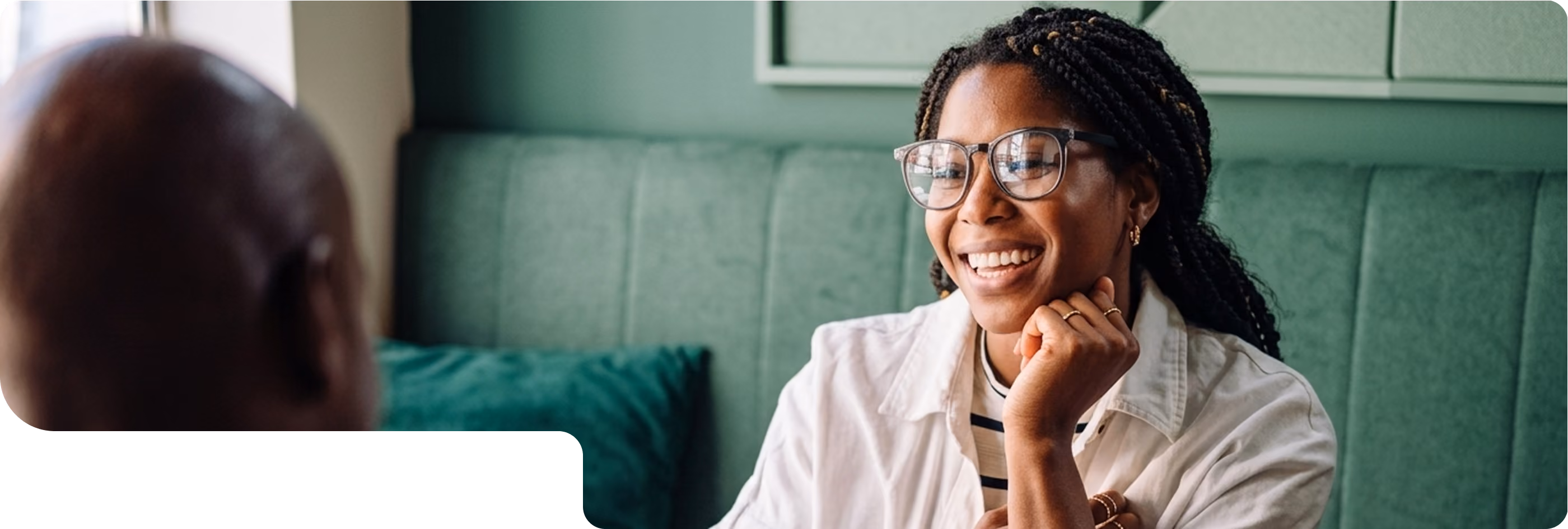 Smiling woman with glasses and braided hair sitting indoors, facing a person whose back is to the camera.