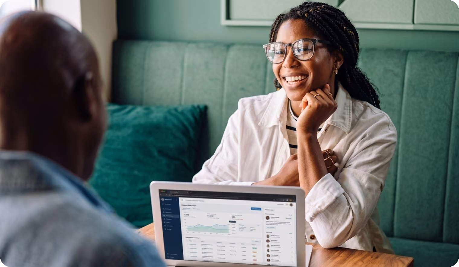 A woman with glasses and braided hair smiles while having a conversation with a man, with a laptop open on the table displaying data charts and lists.