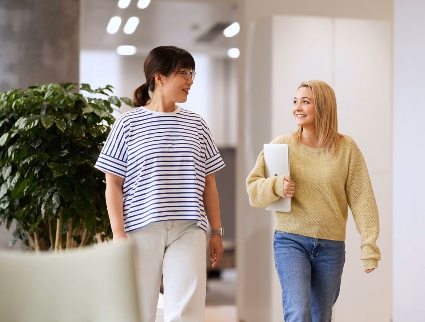 Two women walking and talking in a bright office hallway, one wearing a striped shirt and glasses, the other holding a laptop and wearing a yellow sweater.