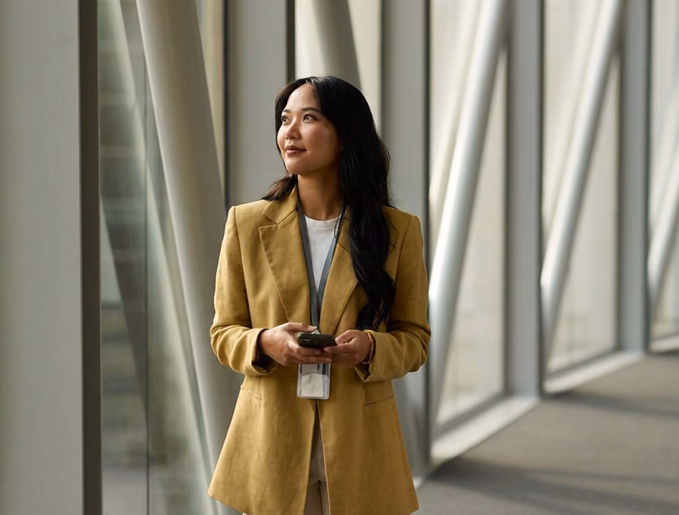 A woman wearing a mustard yellow blazer and holding a phone stands inside a modern building with large windows and structural beams, looking up thoughtfully.