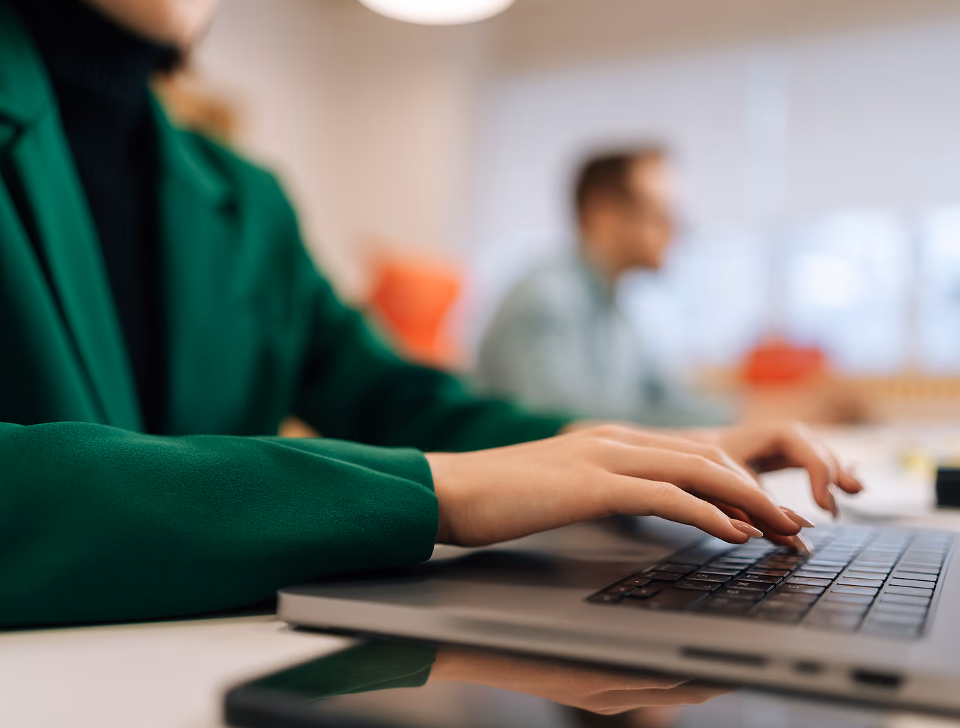 Person in a green jacket typing on a laptop keyboard with another person blurred in the background in an office setting.
