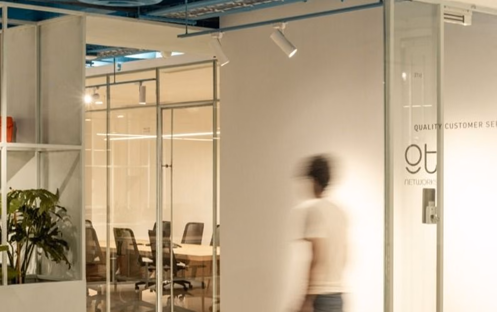 Office interior with glass-walled conference rooms, black office chairs, a plant on a white shelf, and a blurred person walking past.