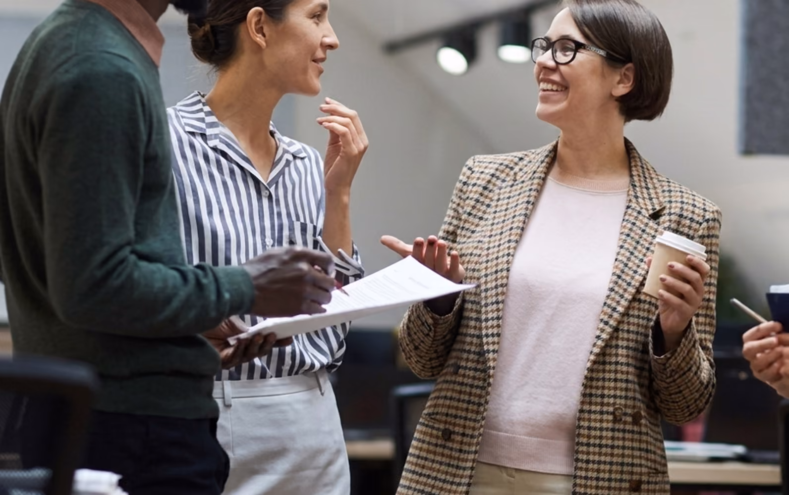 Three colleagues engaged in a friendly discussion in an office; one woman in glasses and a plaid blazer holds a coffee cup while smiling, another woman in a striped shirt listens attentively, and a man holds a clipboard with documents.