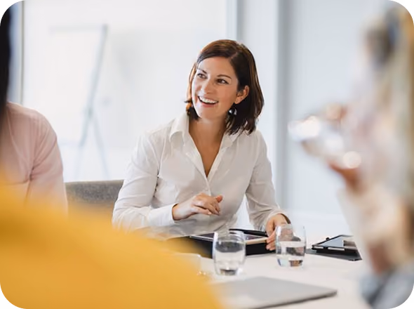 Smiling woman in a white shirt engaged in conversation at a meeting table with glasses of water and notebooks.