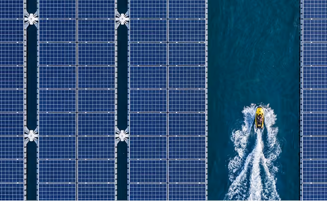 Aerial view of a yellow jet ski creating a white water trail as it moves through blue water between rows of floating blue solar panels.