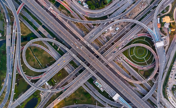 Aerial view of a complex highway interchange with multiple curving ramps, overpasses, and roads crossing over green spaces and buildings.