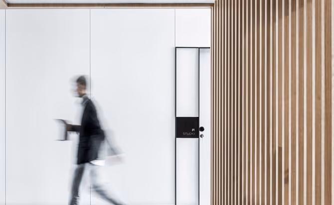 Blurred motion image of a person walking past a modern white wall with a door labeled 'STUDIO' and wooden vertical slats on the right.