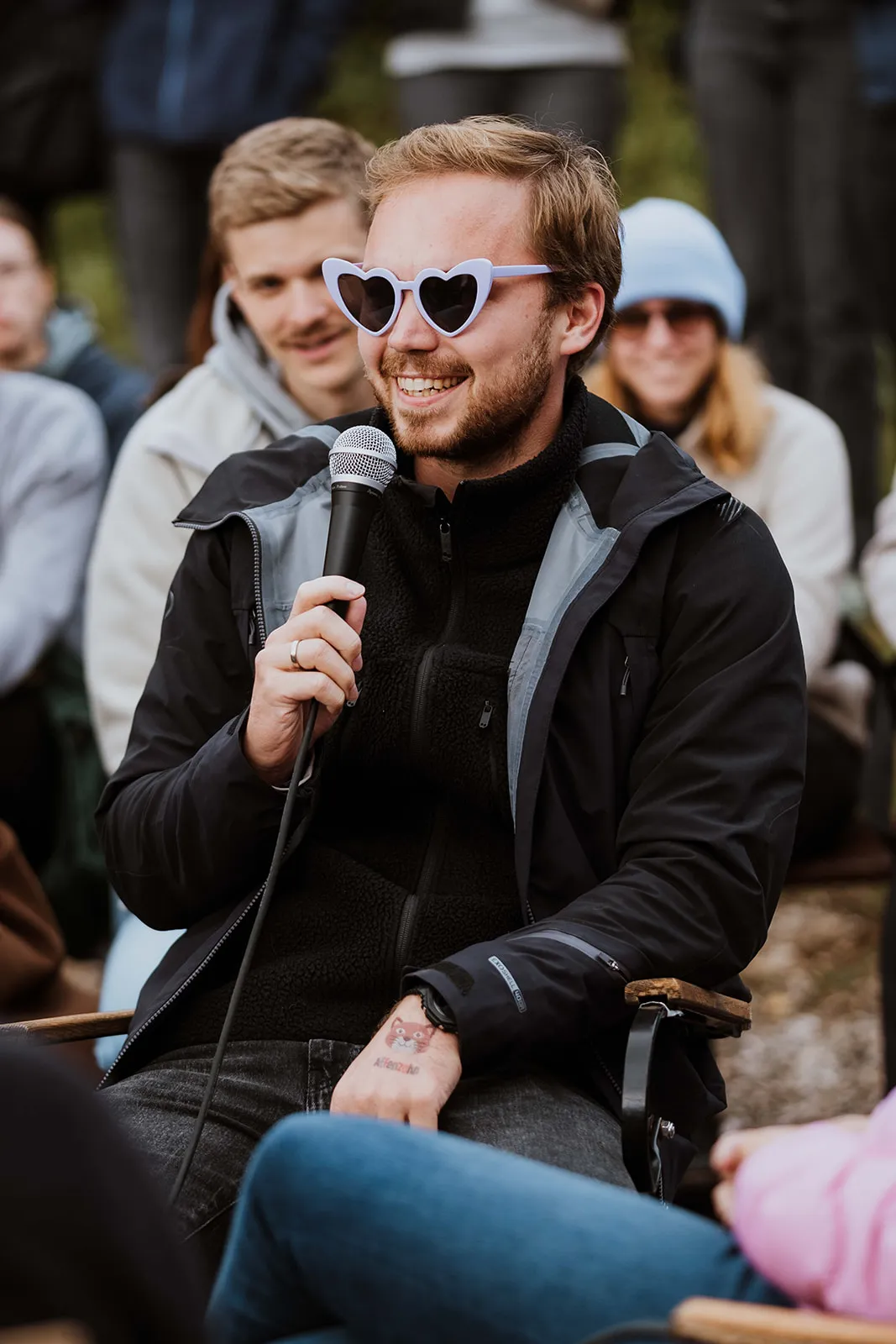 Mann mit herzförmiger Sonnenbrille hält Mikrofon und lächelt, während andere im Hintergrund sitzen.