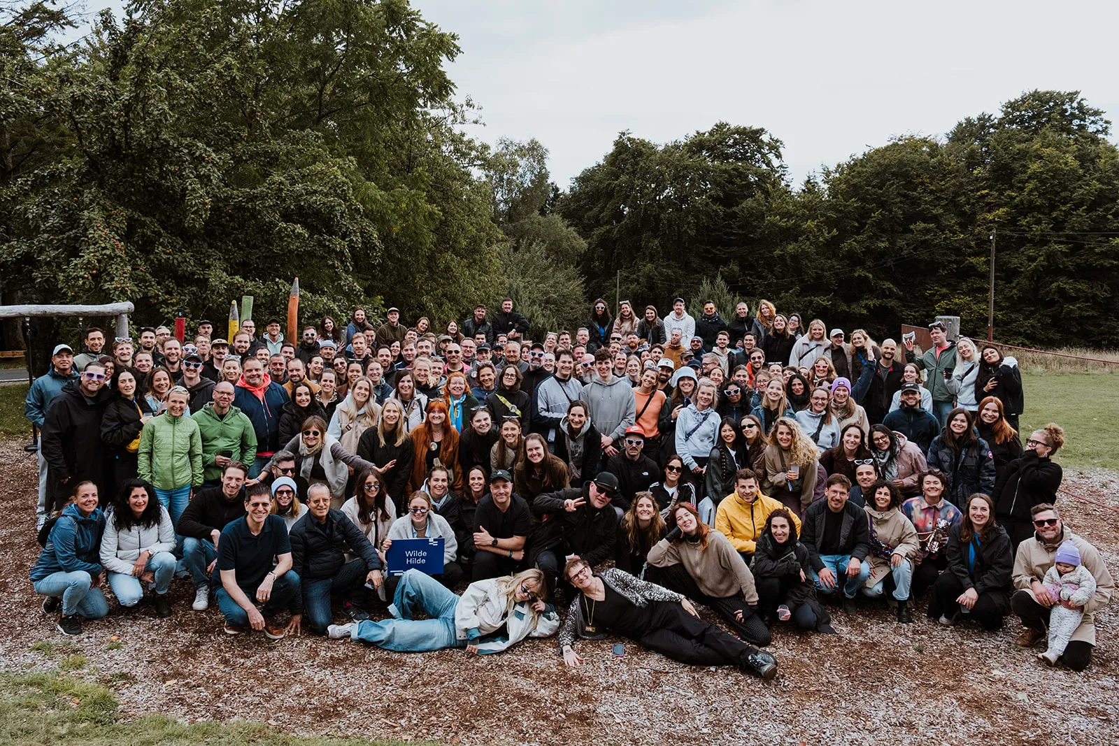 Große Gruppe von Menschen in einem Park vor Bäumen, einige sitzen, andere stehen und lächeln.