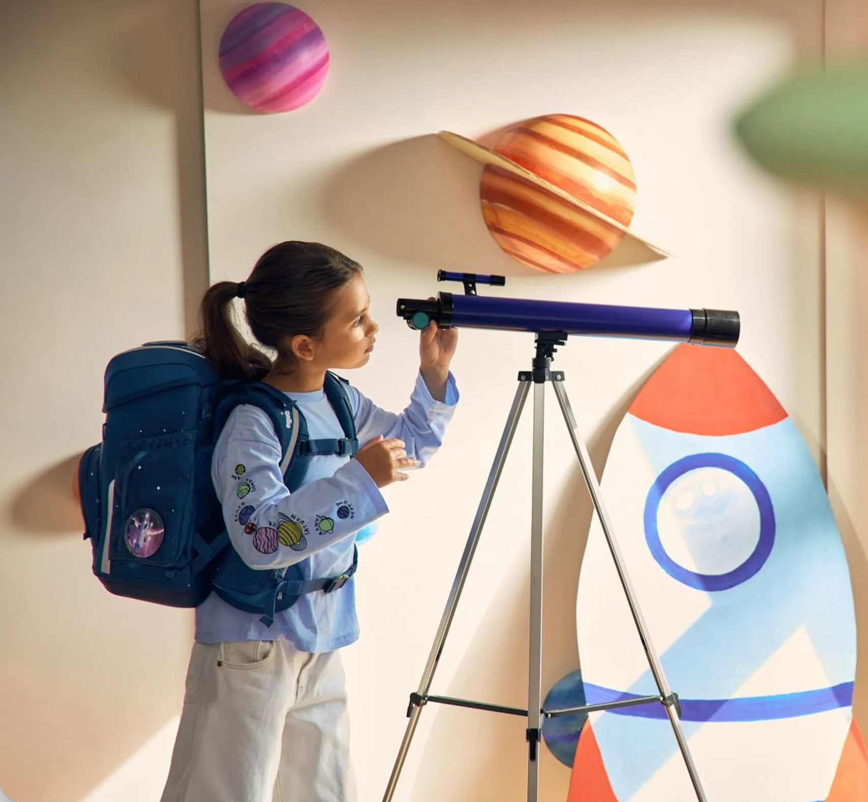 Mädchen mit Weltraum-Rucksack schaut durch Teleskop vor Wand mit Planeten und Rakete Dekoration.