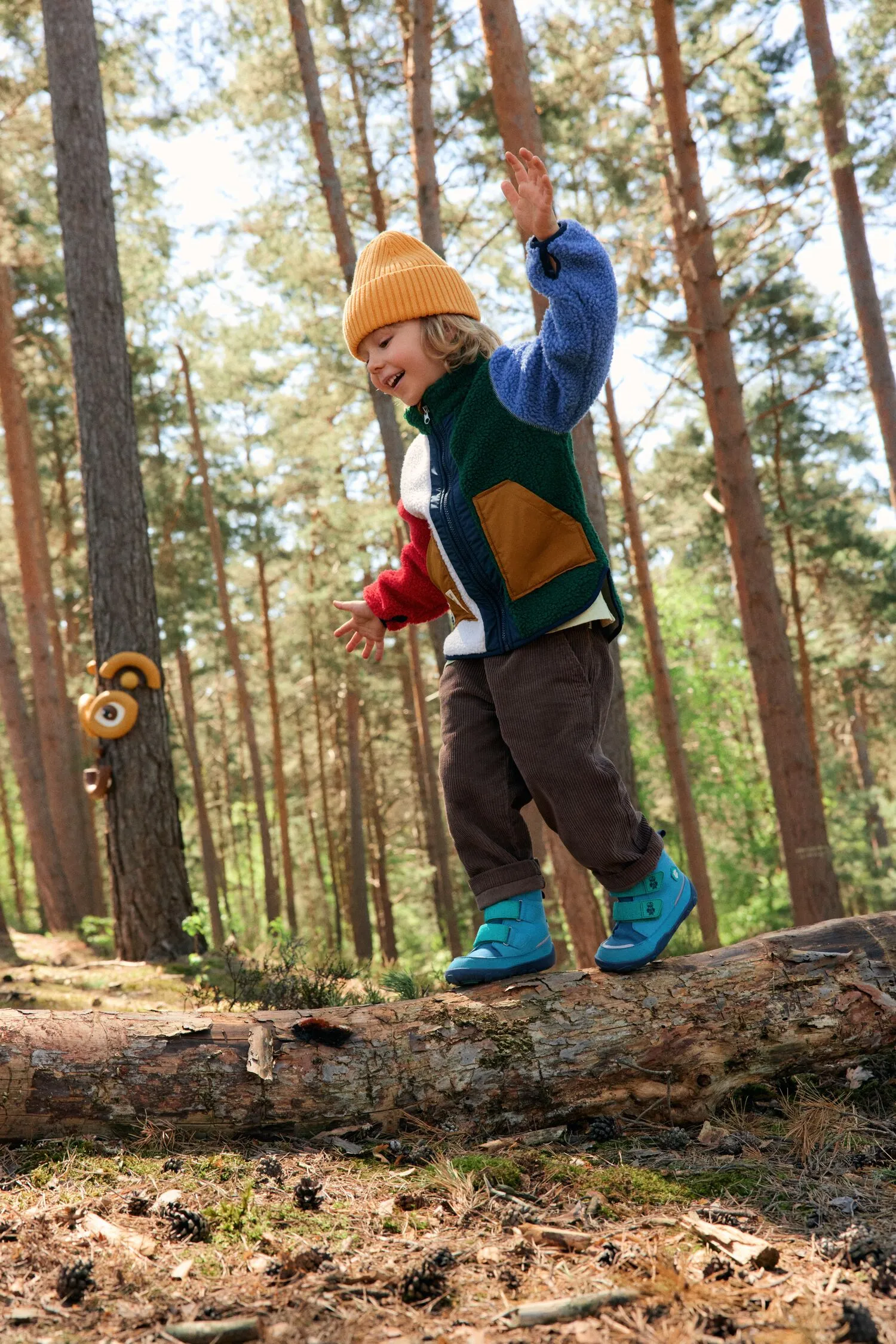 Kind balanciert auf einem liegenden Baumstamm im Wald, trägt bunte Jacke und orangefarbene Mütze.
