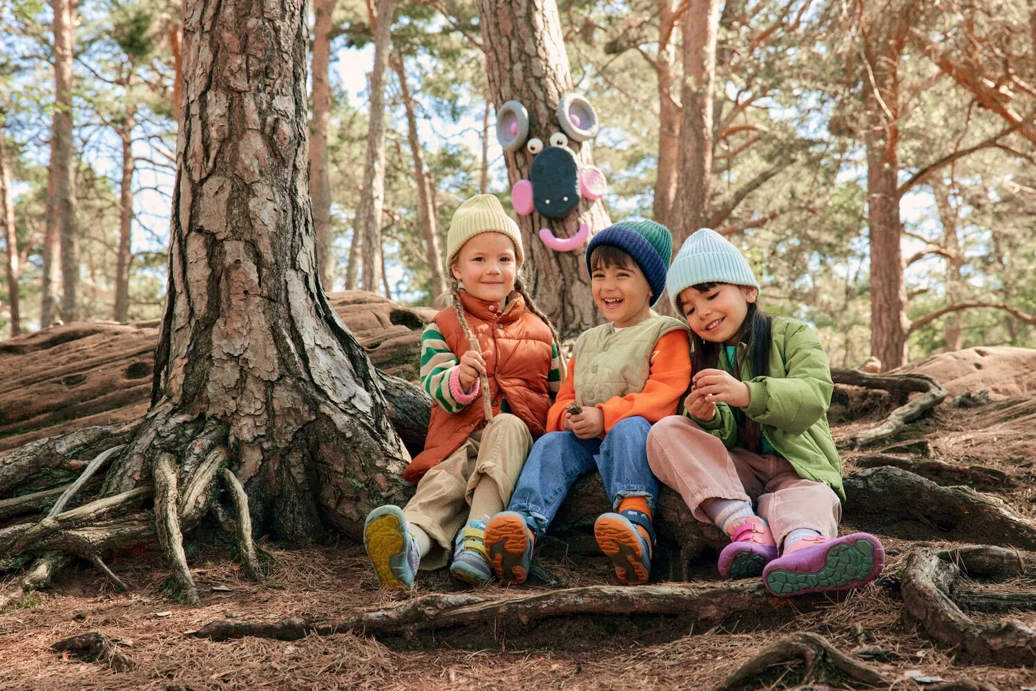 Drei Kinder in bunter Herbstkleidung sitzen lächelnd im Wald vor einem Baum mit einem Gesicht aus Spielzeug.