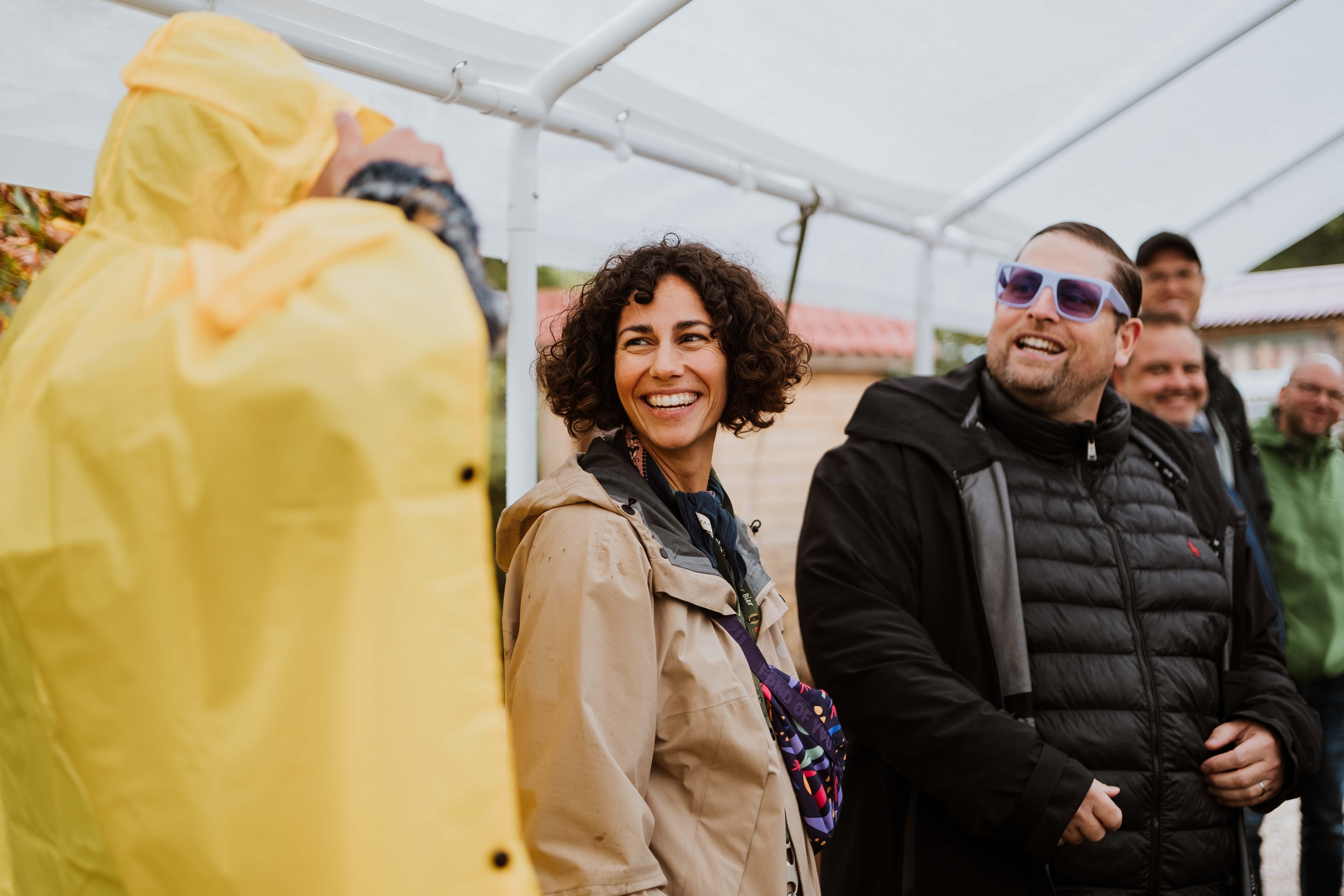 Gruppe von Menschen lacht unter einem weißen Dach, eine Frau trägt beige Jacke, ein Mann Sonnenbrille.