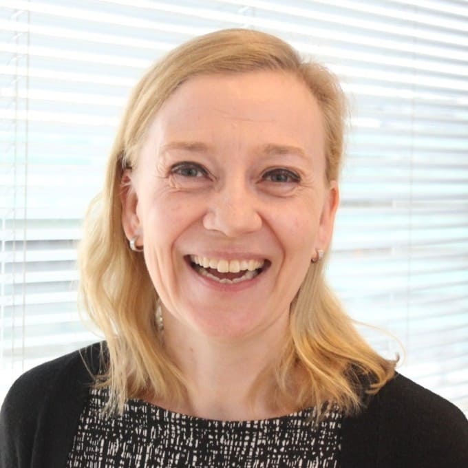 Smiling woman with blonde hair wearing earrings and a black and white top, standing before white blinds.