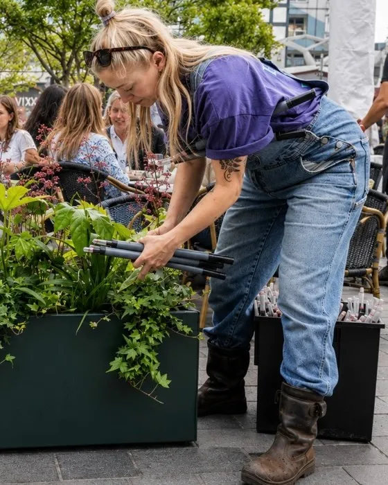 Medewerker in paars shirt verzorgt een donkergroene plantenbak op het terras van Noord