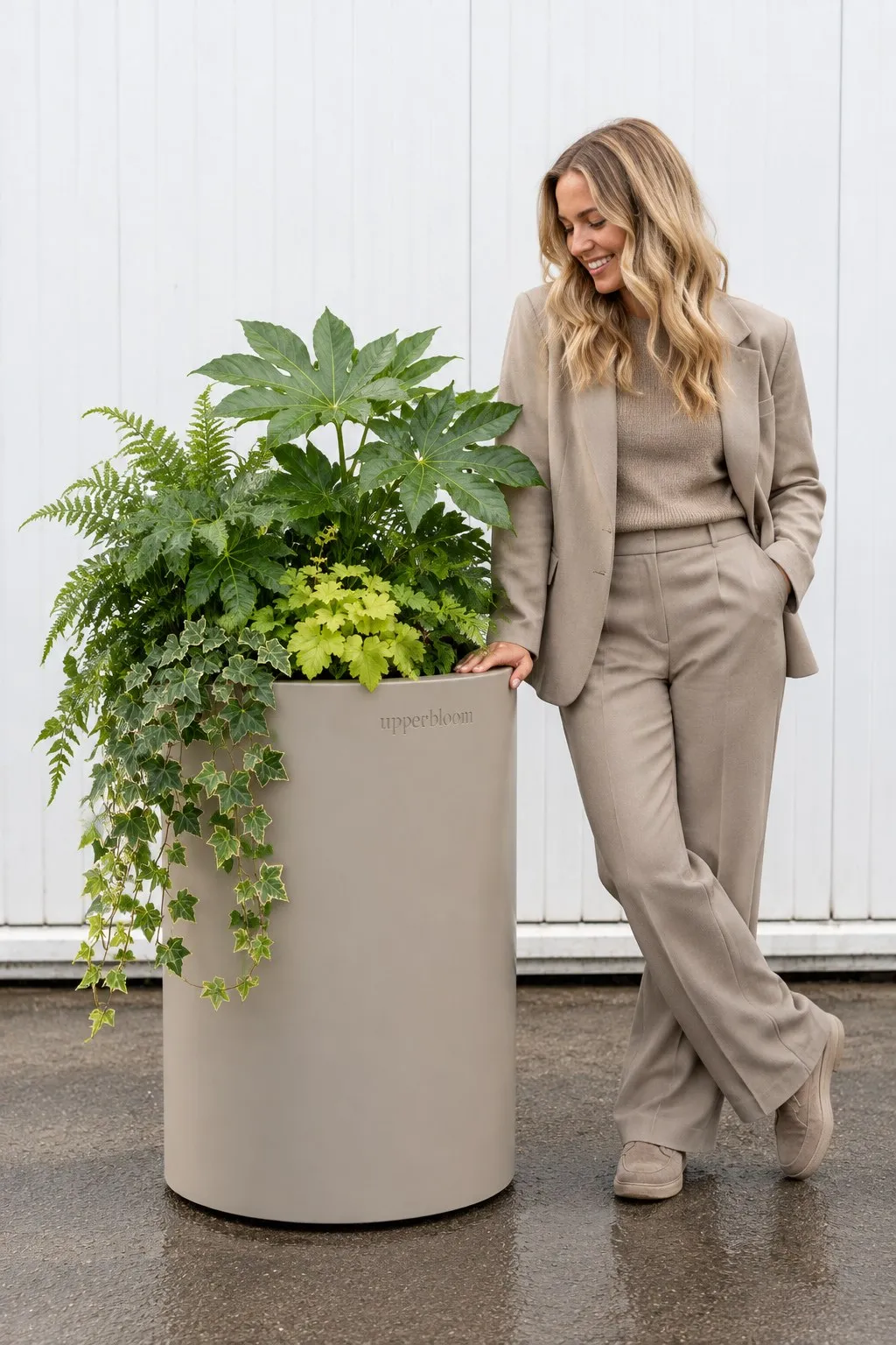 Vrouw in beige pak leunt op een hoge ronde taupe plantenbak met varens en klimop