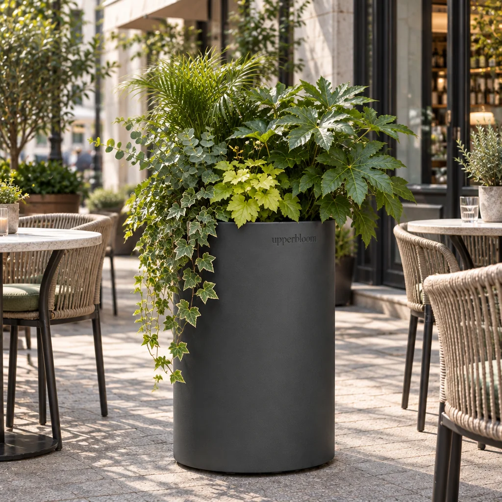 Ronde antraciet plantenbak met varens, vingerplant en klimop op een terras met rieten stoelen