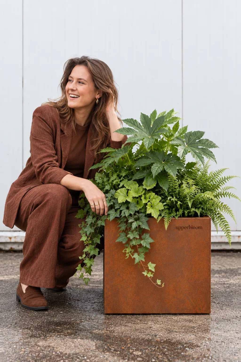Lachende vrouw geknield naast een cortenstalen plantenbak met varens, vingerplant en klimop